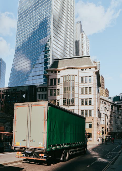 A large green delivery truck parked on a city street with modern high-rise buildings and older architecture in the background. The truck's cargo area is closed, and it is positioned near the pavement,
