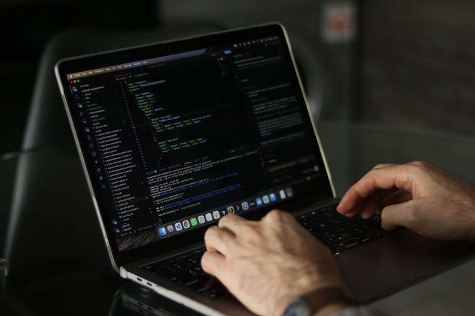 A person with dark hair, wearing a light blue shirt, is seated at a desk during a home relocation process, working on a laptop displaying a screen filled with colorful lines of code. The individual is